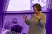 Photograph of Mary Kay Chess explaining something to the class with her hands in front of her and a screen behind