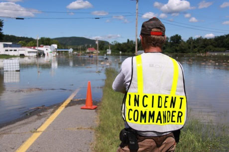 Photograph of a man in a vest reading Incident Commander looking at a flooded road.