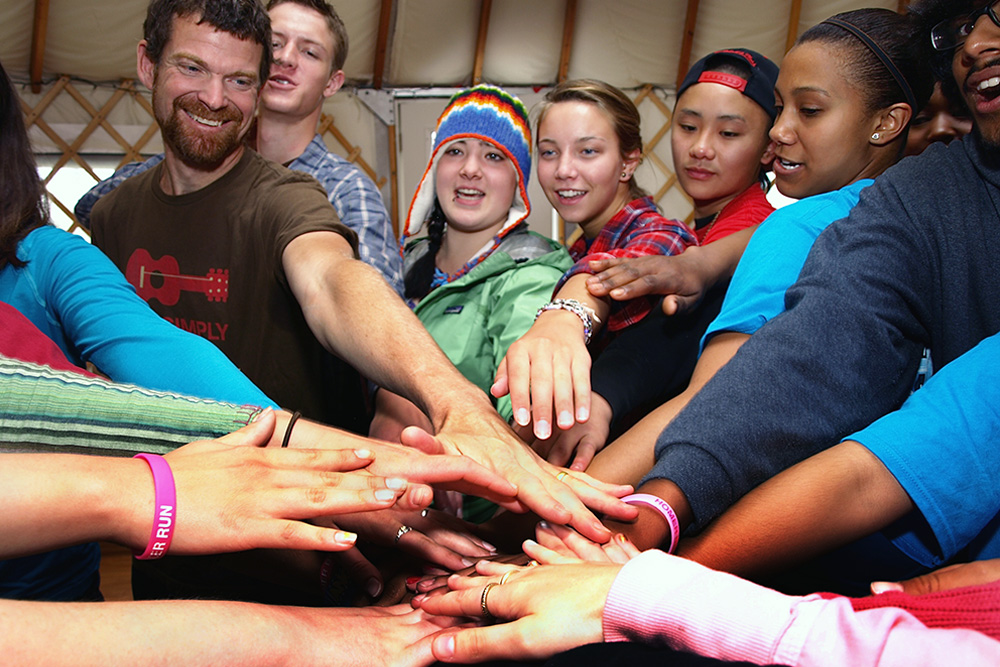 Photograph of several teens clasping hands in the center of a circle.