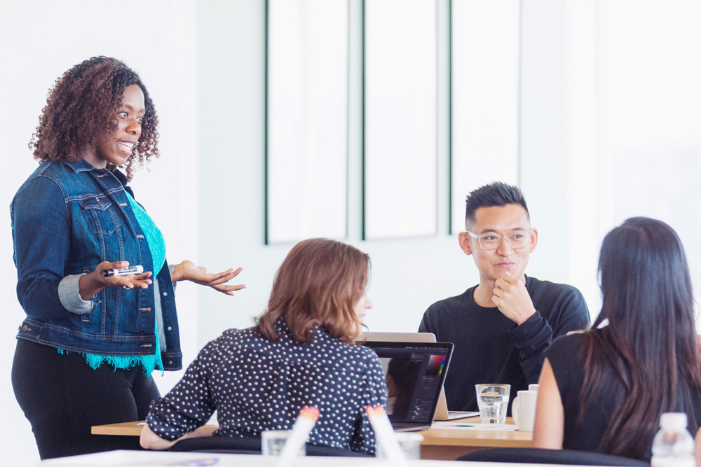 Photograph of a person talking to people seated around a table.