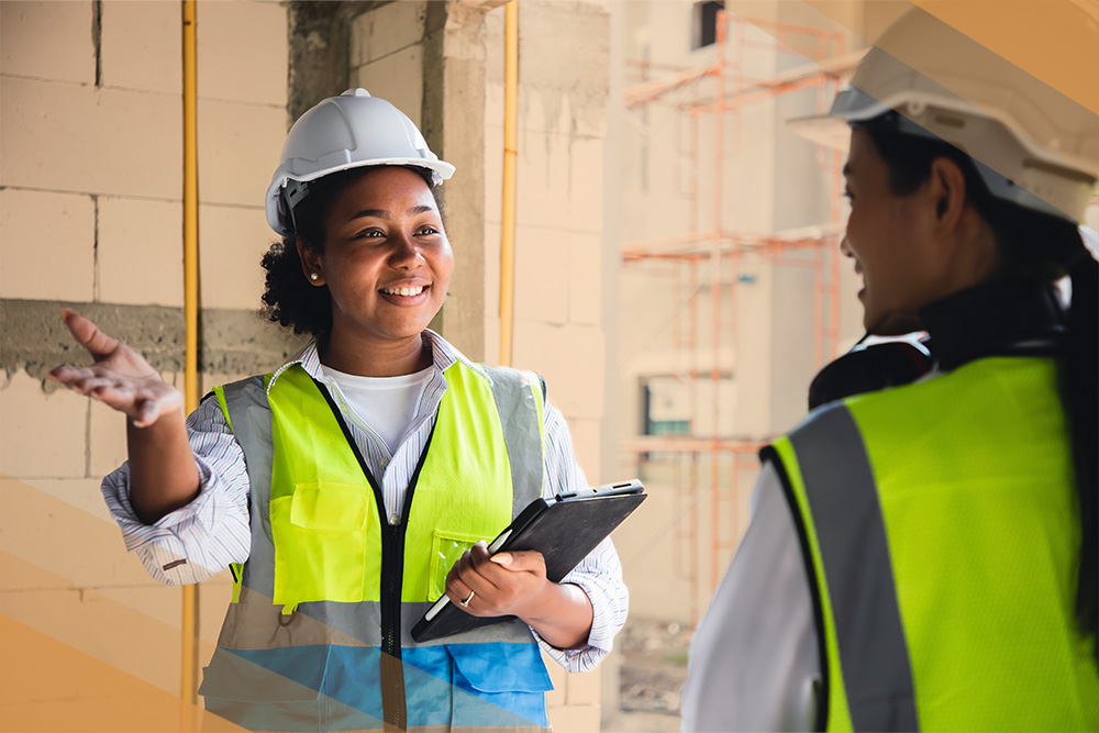 Image of two people with protective vests and hardhats talking with each other. One holds a clipboard.